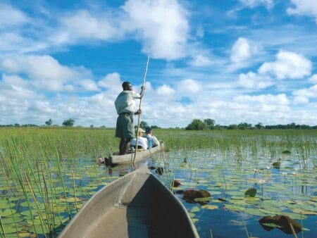 Drei Personen in einem Einbaum-Boot auf dem Wasser umgeben von Seerosen im Okavango Delta in Botswana.